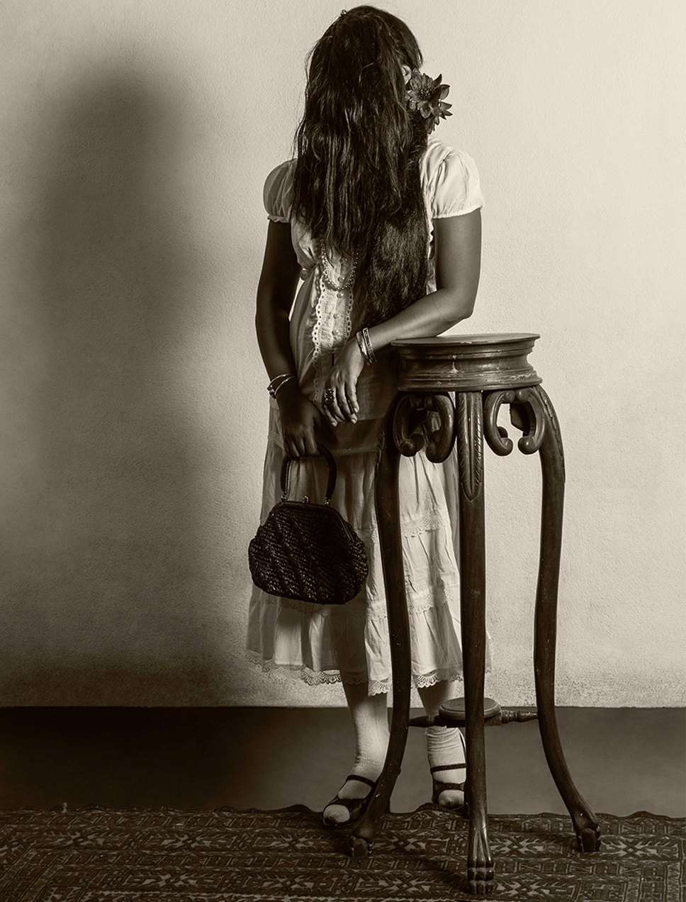 At the centre is a woman whose face isn’t visible as it is completely covered by her long hair. A single flower is tucked behind her ear. She wears a dress, carries a small handbag in one hand and rests the other on a tall wooden table in front of her.