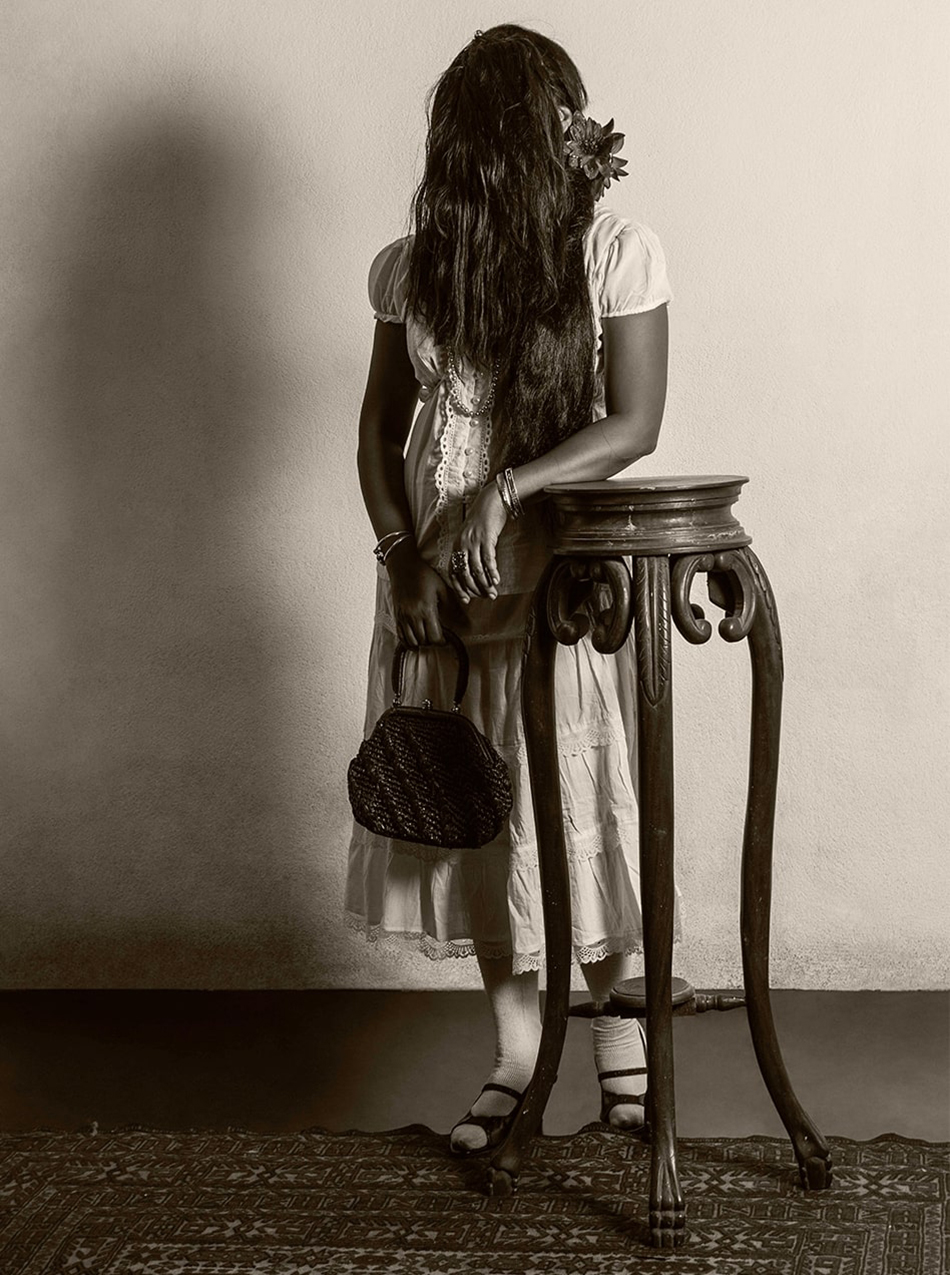 At the centre is a woman whose face isn’t visible as it is completely covered by her long hair. A single flower is tucked behind her ear. She wears a dress, carries a small handbag in one hand and rests the other on a tall wooden table in front of her.