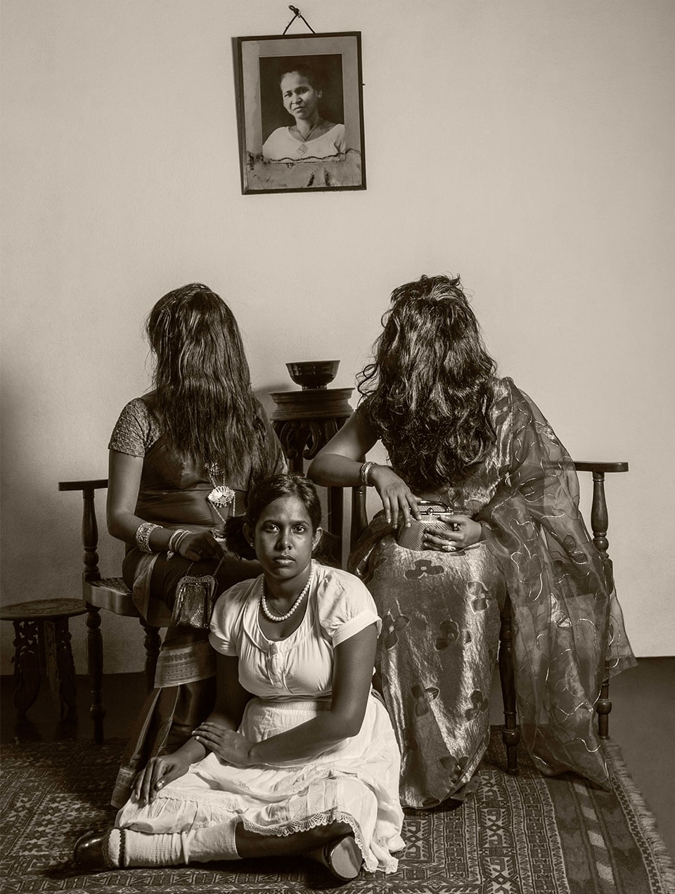 A woman in a dress sits cross legged on a carpet. Behind her are two saree-clad women seated on chairs, with their hair completely covering their faces. 