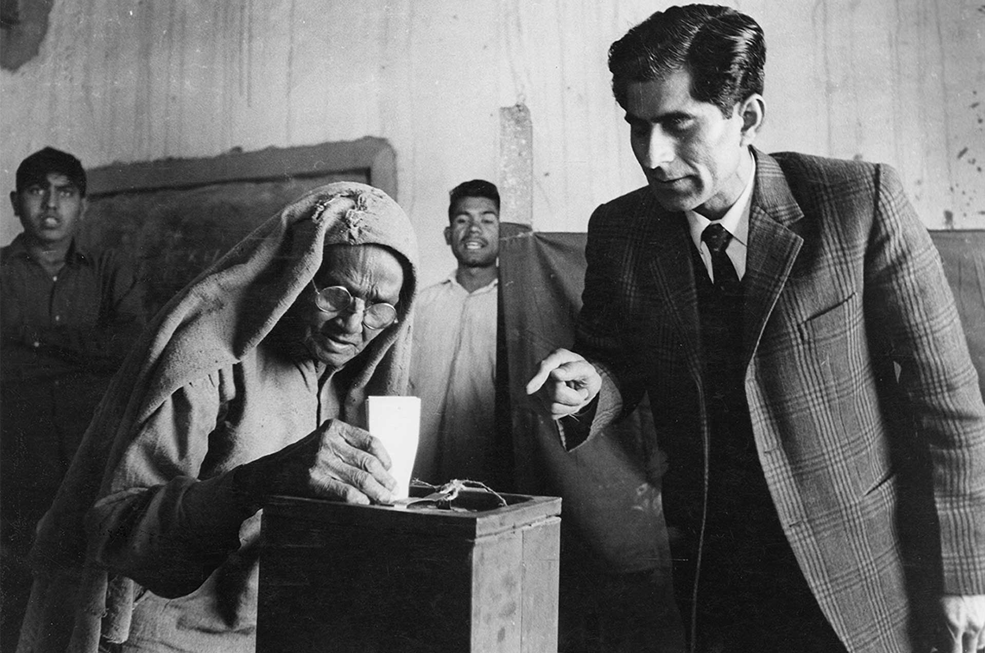 A bespectacled old woman slips a paper into a ballot box, as a middle-aged man in a suit points at it. There are two men in the background. One looks on, while the other looks elsewhere. 