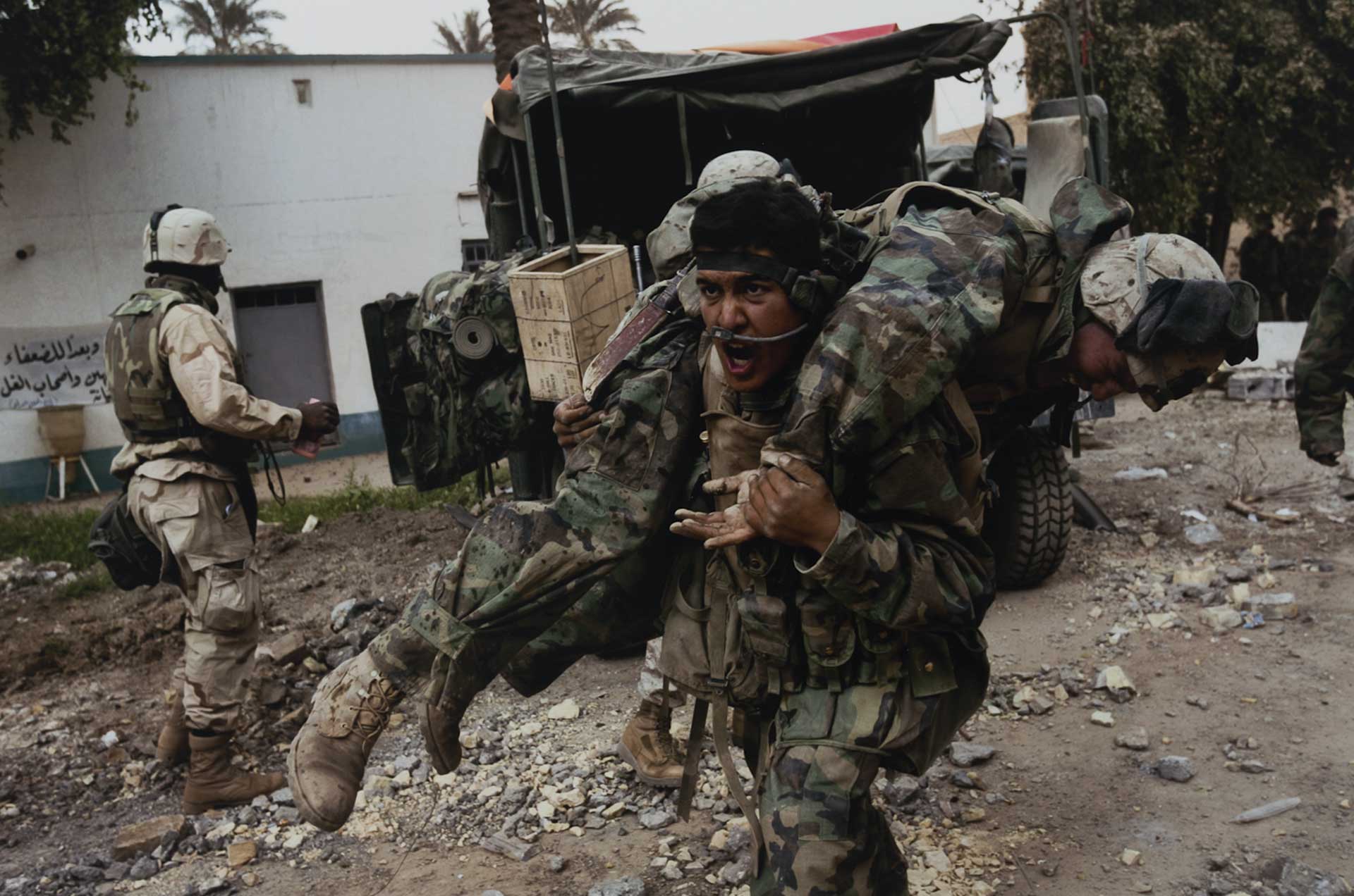 A screaming soldier carrying another soldier across his shoulders as he charges forward. In the background, a man stands next to a truck. All the men wear combat clothes. 