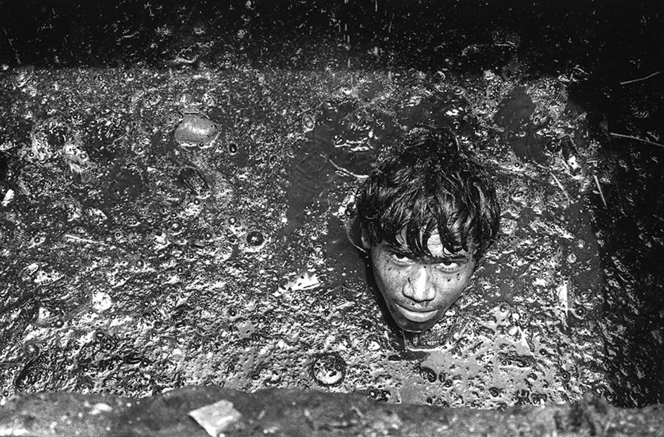The head of a young man, as if floating, surrounded by murky sewage water. He seems to be looking up.