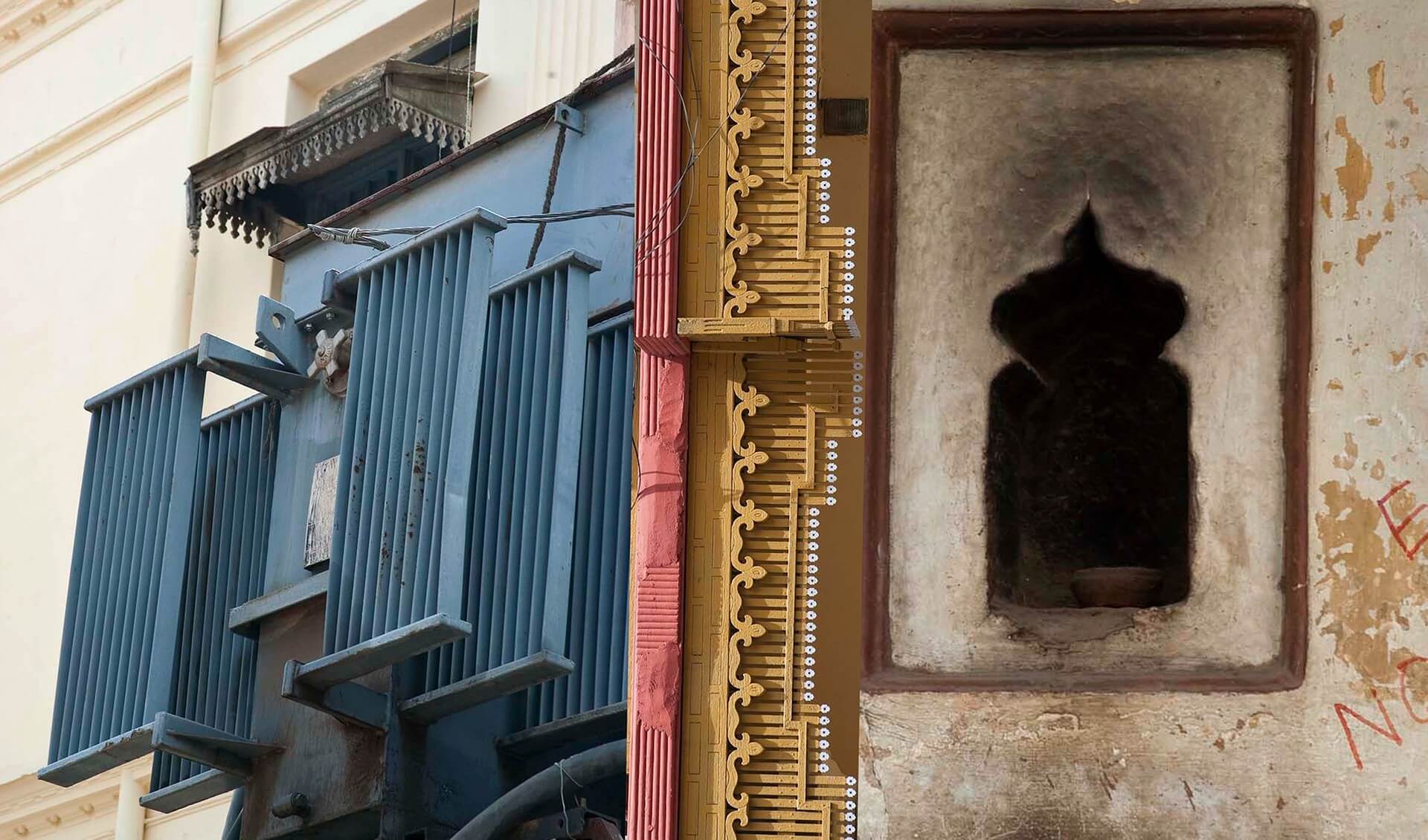 Blue transformer mounted on a window. Next to it is pink and yellow geometric wall design and a pale dark space with a hazy lamp or diya inside it.