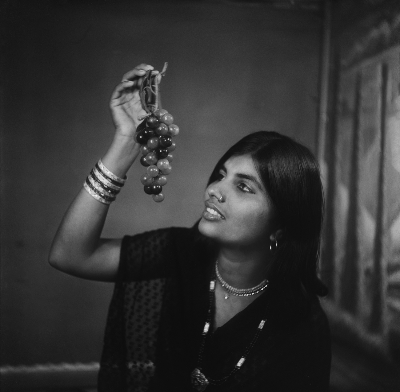 Woman wearing a black saree looking towards a bunch of grapes held in her left hand. She is wearing bangles on her left hand and a necklace.