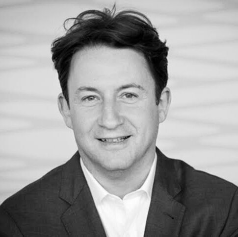 Andras Szanto smiling and looking directly at the camera in black and white wearing a collared shirt and lapelled jacket, his hair slightly tousled.