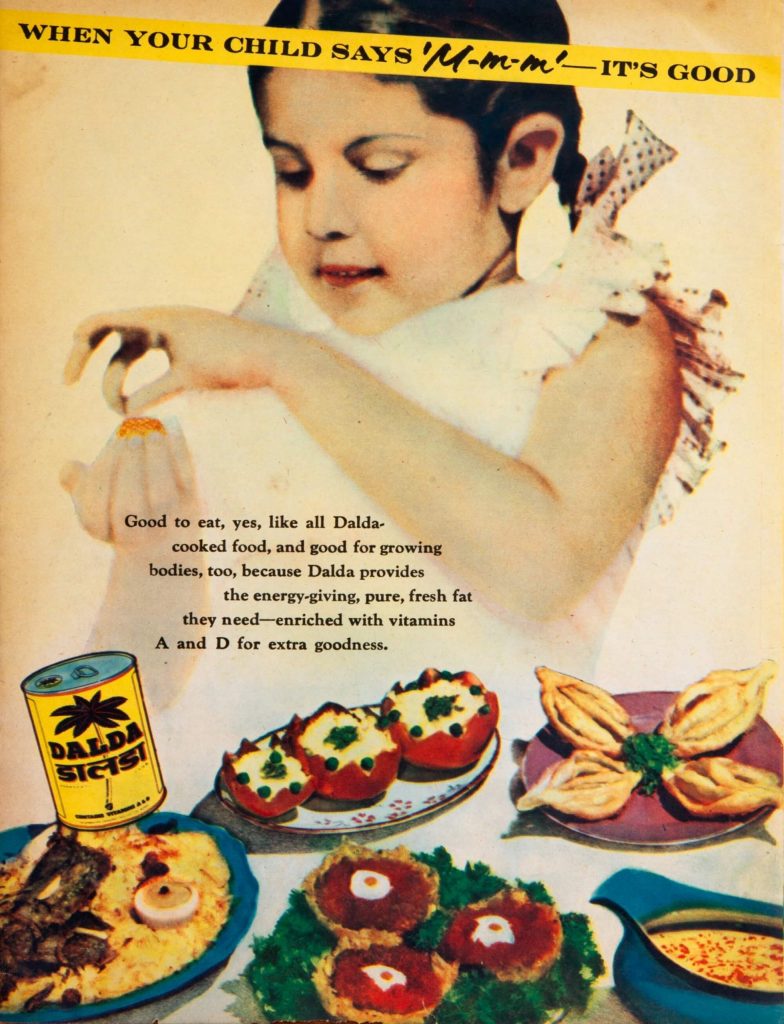 A young girl eagerly peels the wrapper of a laddoo like dessert. Below her is a tin of dalda and a table full of sweets like gujiya.