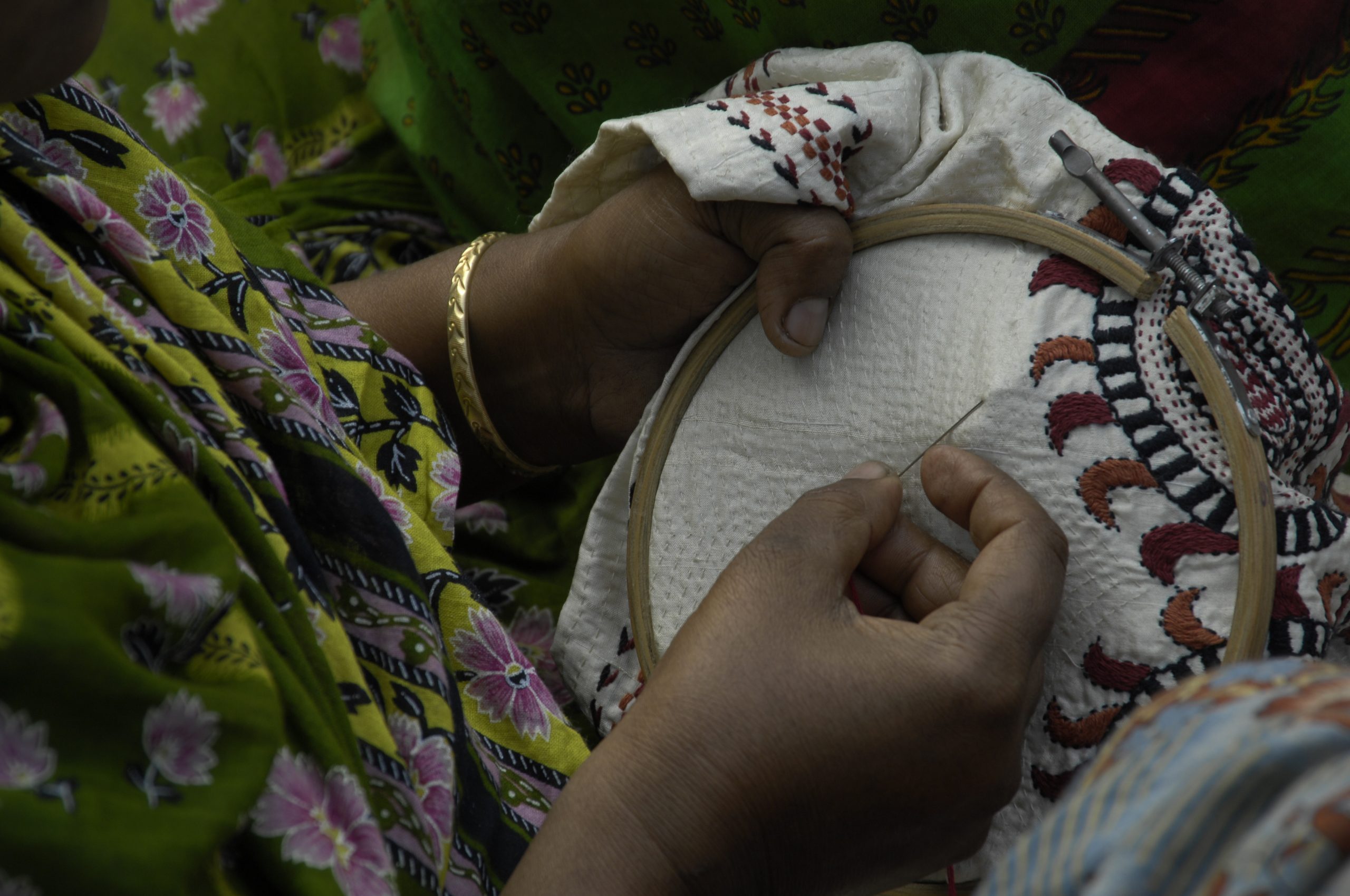 Hands holding an embroidery hoop a needle in right hand is stitching shades of brown onto a white. Golden bangle on left wrist. Wearing a green outfit printed with flowers.